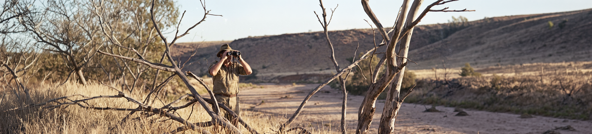 Hunter in khaki using binoculars among dead trees in a dry outback creek bed