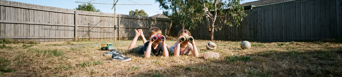 Two kids lying on the backyard lawn looking through colourful kids binoculars