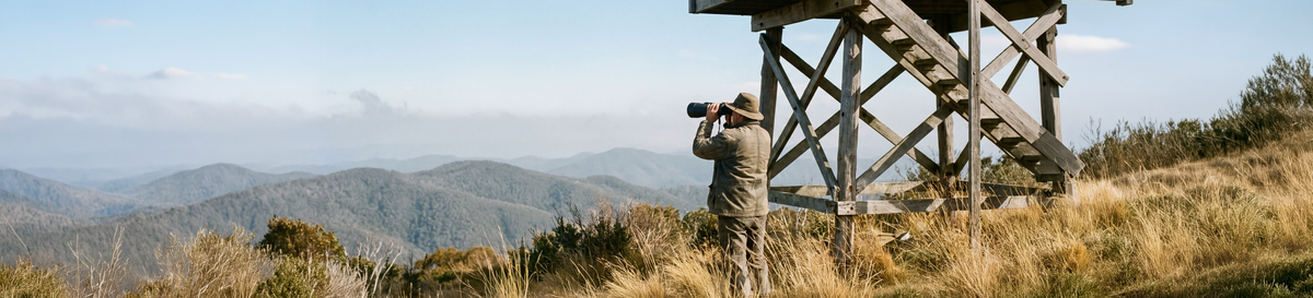 Man using large professional binoculars beside a timber lookout tower in the mountains